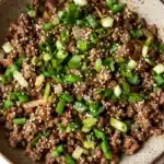 Korean beef skillet - the image is a close-up of a bowl of food. the bowl is made of ceramic and has a brown rim. the food appears to be a dish made with ground beef, sliced green onions, and sesame seeds. the ground beef is dark brown and looks crispy on the outside. the onions are bright green and are scattered throughout the dish. the sesame seed is visible on top of the ground beef. the dish is garnished with a sprinkle of sesame oil. the background is a white marble countertop.