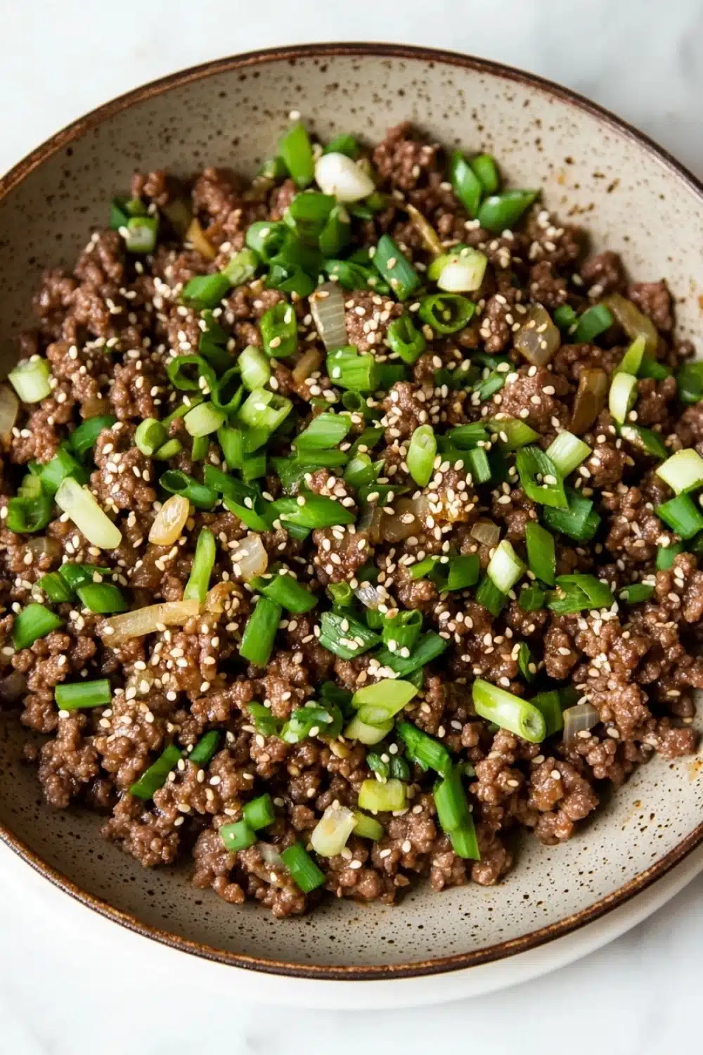 Korean beef skillet - the image is a close-up of a bowl of food. the bowl is made of ceramic and has a brown rim. the food appears to be a dish made with ground beef, sliced green onions, and sesame seeds. the ground beef is dark brown and looks crispy on the outside. the onions are bright green and are scattered throughout the dish. the sesame seed is visible on top of the ground beef. the dish is garnished with a sprinkle of sesame oil. the background is a white marble countertop.