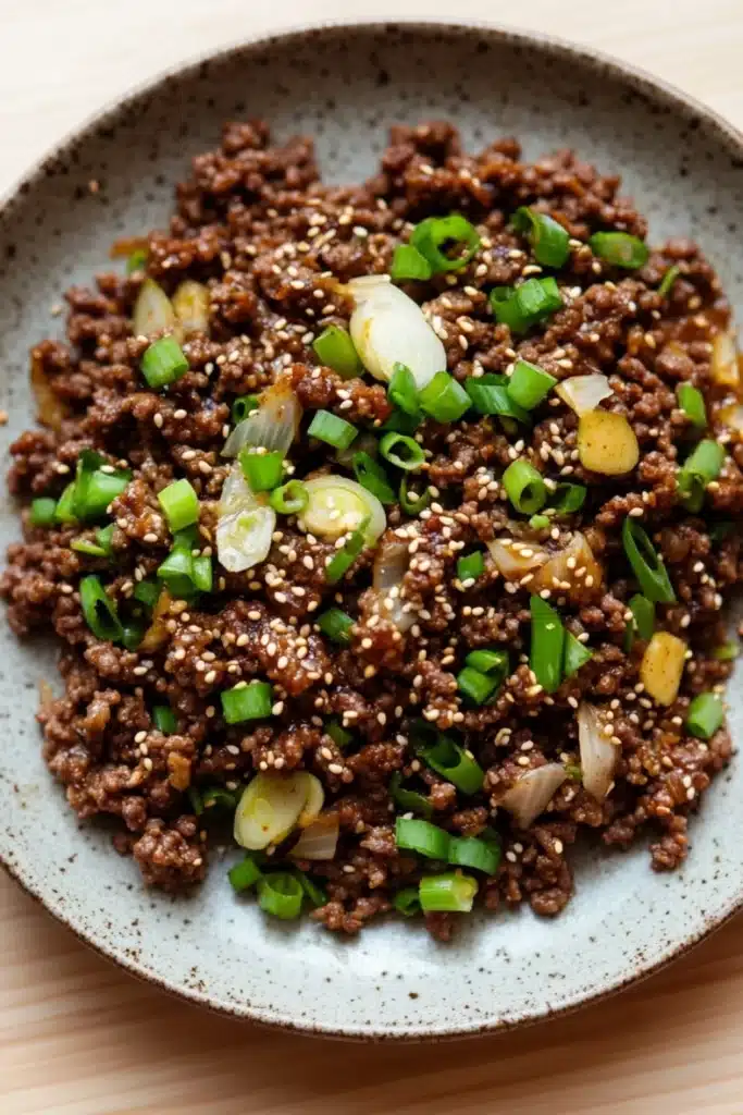 Korean beef skillet - the image is a close-up of a bowl of food. the bowl is made of a light-colored ceramic with a speckled pattern. the food appears to be a dish made with ground beef, sliced green onions, and small pieces of yellow squash. the ground beef is dark brown and looks crispy on the outside. the onions are cut into small cubes and are scattered throughout the dish. the green onions are bright green and look fresh and vibrant. the yellow squash is cut into thin slices and is placed on top of the ground beef. the dish is garnished with sesame seeds, giving it a crunchy texture.