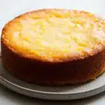 Lemon cake layer - the image is of a round cake on a white plate. the cake appears to be freshly baked and has a golden brown crust on top. the top of the cake is covered in a layer of yellow sugar, which is sprinkled with small orange zest. the plate is round and white, and the cake looks moist and fluffy. the background is a plain white surface.