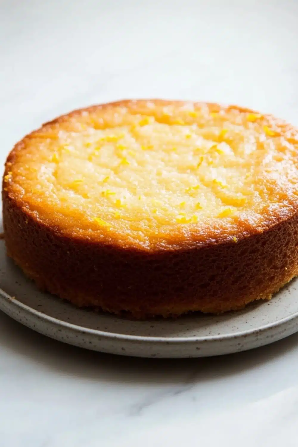 Lemon cake layer - the image is of a round cake on a white plate. the cake appears to be freshly baked and has a golden brown crust on top. the top of the cake is covered in a layer of yellow sugar, which is sprinkled with small orange zest. the plate is round and white, and the cake looks moist and fluffy. the background is a plain white surface.