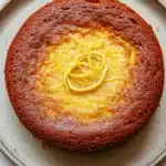 Lemon cake low sugar - the image is a close-up of a round cake on a white plate. the cake appears to be a chocolate cake with a golden brown crust on top. the top of the cake is covered in a layer of orange-colored frosting, which is drizzled with a yellow lemon zest. the frosting is smooth and shiny, and the cake looks moist and fluffy. the plate is round and has a light blue rim. the background is a white marble countertop.