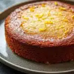 Lemon cake low sugar - the image is a close-up of a round cake on a gray plate. the cake appears to be freshly baked and has a golden brown crust on top. the top of the cake is covered in a light dusting of powdered sugar, giving it a shiny appearance. there are small yellow zigzag lines scattered across the cake, adding a pop of color to the overall appearance. the plate is sitting on a dark, textured surface, possibly a table or countertop.
