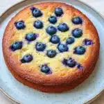 Lemon cake with blueberries - the image is of a round cake on a blue and white plate. the cake appears to be freshly baked and has a golden brown crust on top. on top of the cake, there are small blueberries arranged in a circular pattern. the blueberries are bright blue and appear to be ripe and juicy. the plate is sitting on a light-colored tablecloth.