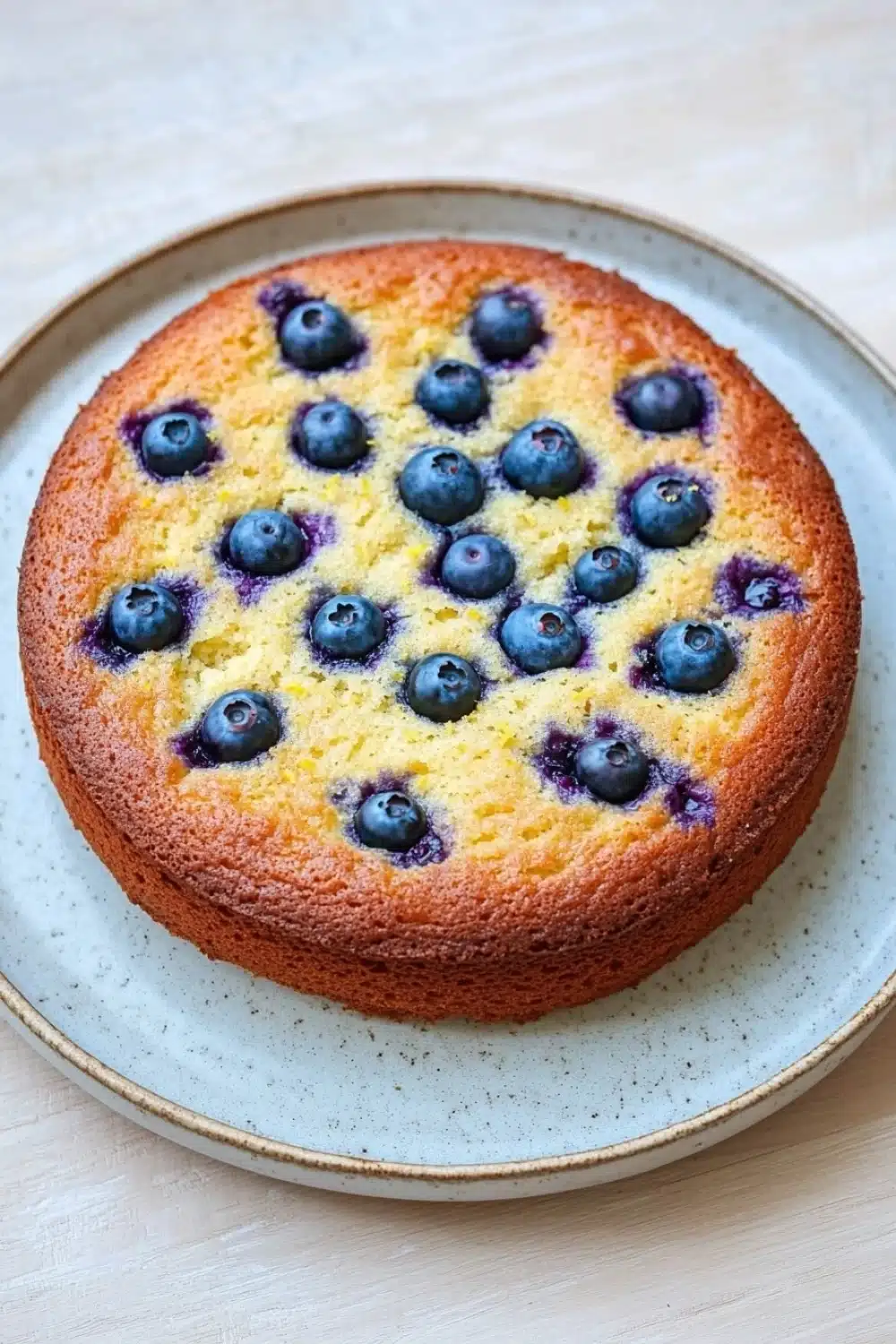 Lemon cake with blueberries - the image is of a round cake on a blue and white plate. the cake appears to be freshly baked and has a golden brown crust on top. on top of the cake, there are small blueberries arranged in a circular pattern. the blueberries are bright blue and appear to be ripe and juicy. the plate is sitting on a light-colored tablecloth.
