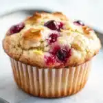 Lemon cherry muffin - the image is of a freshly baked muffin on a white marble countertop. the muffin is golden brown in color and has a crumbly texture. it is topped with fresh cranberries and lemon zest, giving it a sweet and tart flavor. the background is blurred, making the muffin the focal point of the image.