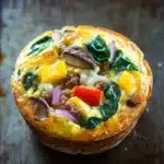 Low cal freezer breakfast - the image shows a small quiche on a baking tray. the quiche is golden brown and has a flaky crust. it is filled with a mixture of scrambled eggs, sausage, red onions, yellow bell peppers, and green spinach leaves. there are also a few mushrooms scattered throughout the mixture. the tray is lined with parchment paper and the background is blurred, making the quiche the focal point of the image.