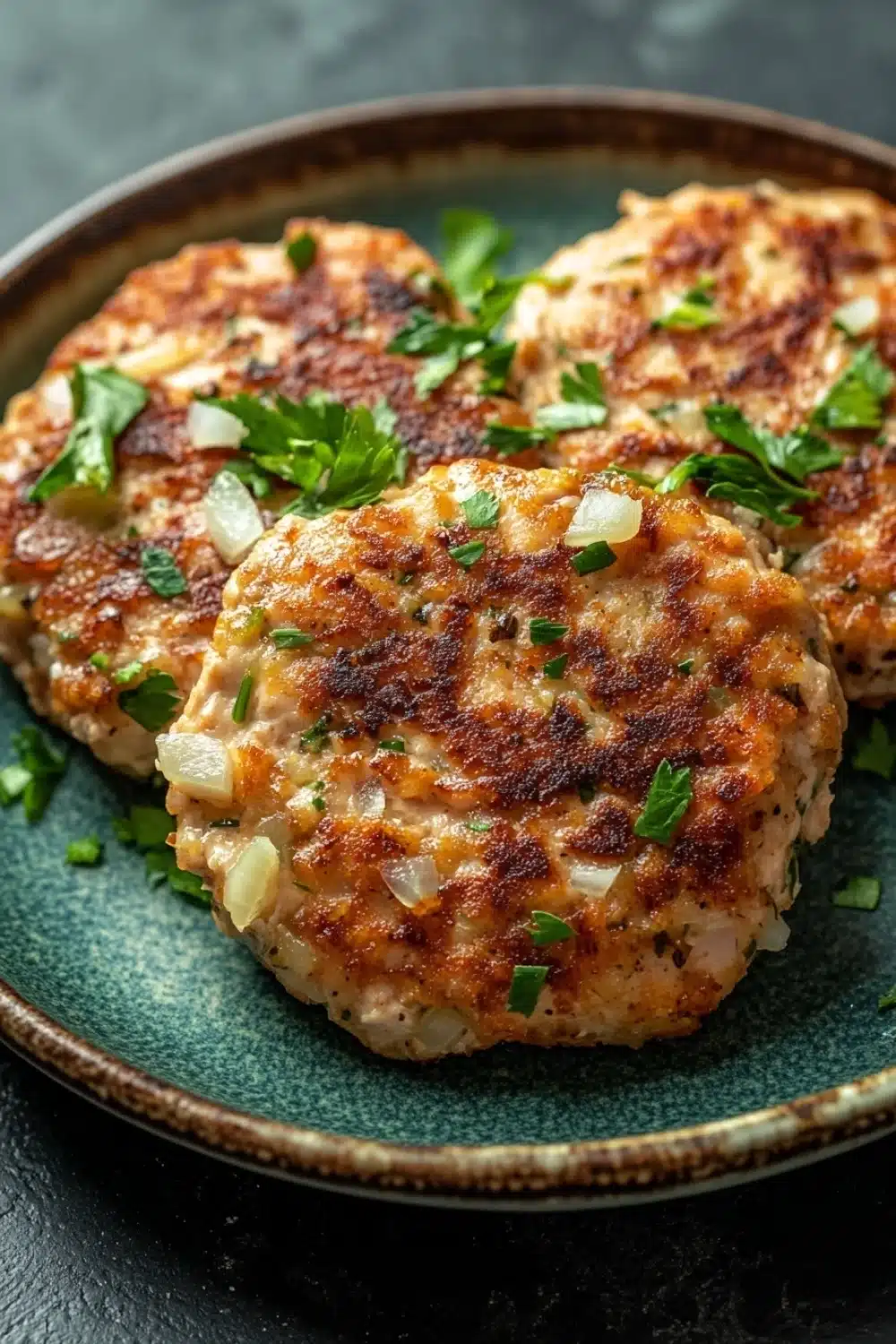 Low calorie chicken patties - the image shows three crab cakes on a blue plate. the crab cakes are golden brown and appear to be seasoned with herbs and spices. they are arranged in a circular pattern on the plate, with some overlapping each other. the plate is garnished with a sprig of fresh parsley. the background is a dark grey countertop.