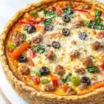 Low carb deep dish pizza quiche - the image is of a freshly baked quiche on a wooden cutting board. the quiche has a golden brown crust and is filled with a variety of ingredients. on top of the quiche, there are sliced red and green bell peppers, black olives, and crumbled cheese. the cheese is melted and bubbly, and there are also some green herbs scattered throughout the dish. the background is a white marble countertop.