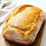 Low carb yeast sandwich bread - the image shows a loaf of bread on a wooden cutting board. the bread appears to be freshly baked and has a golden brown crust on top. the loaf is sitting on a white marble countertop with a white cloth napkin on the side. the background is blurred, making the bread the focal point of the image.