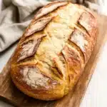 Low carb yeast sandwich bread - the image shows a freshly baked loaf of bread on a wooden cutting board. the bread is golden brown in color and has a criss-cross pattern on the top. it appears to be freshly baked and is sitting on a white marble countertop with a beige cloth napkin next to it. the cutting board is made of light-colored wood and is placed on a flat surface. the background is blurred, so the focus is on the bread.