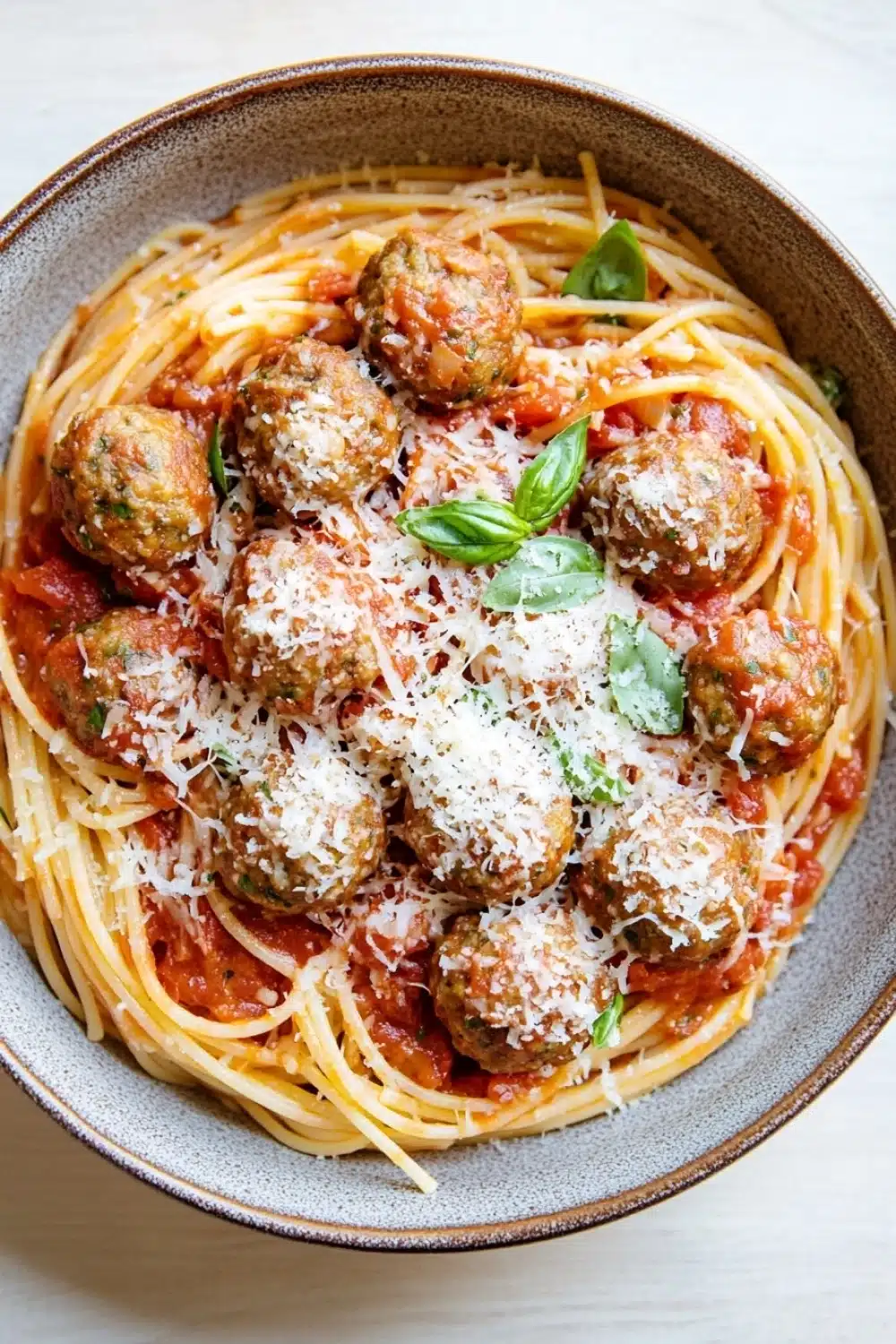 Make ahead pasta and meatballs - the image shows a bowl of spaghetti and meatballs. the spaghetti is cooked al dente and is covered in a red tomato sauce with chunks of meatballs scattered throughout. the meatballs are golden brown and appear to be seasoned with grated parmesan cheese. the dish is garnished with fresh basil leaves. the bowl is made of ceramic and is sitting on a white countertop.