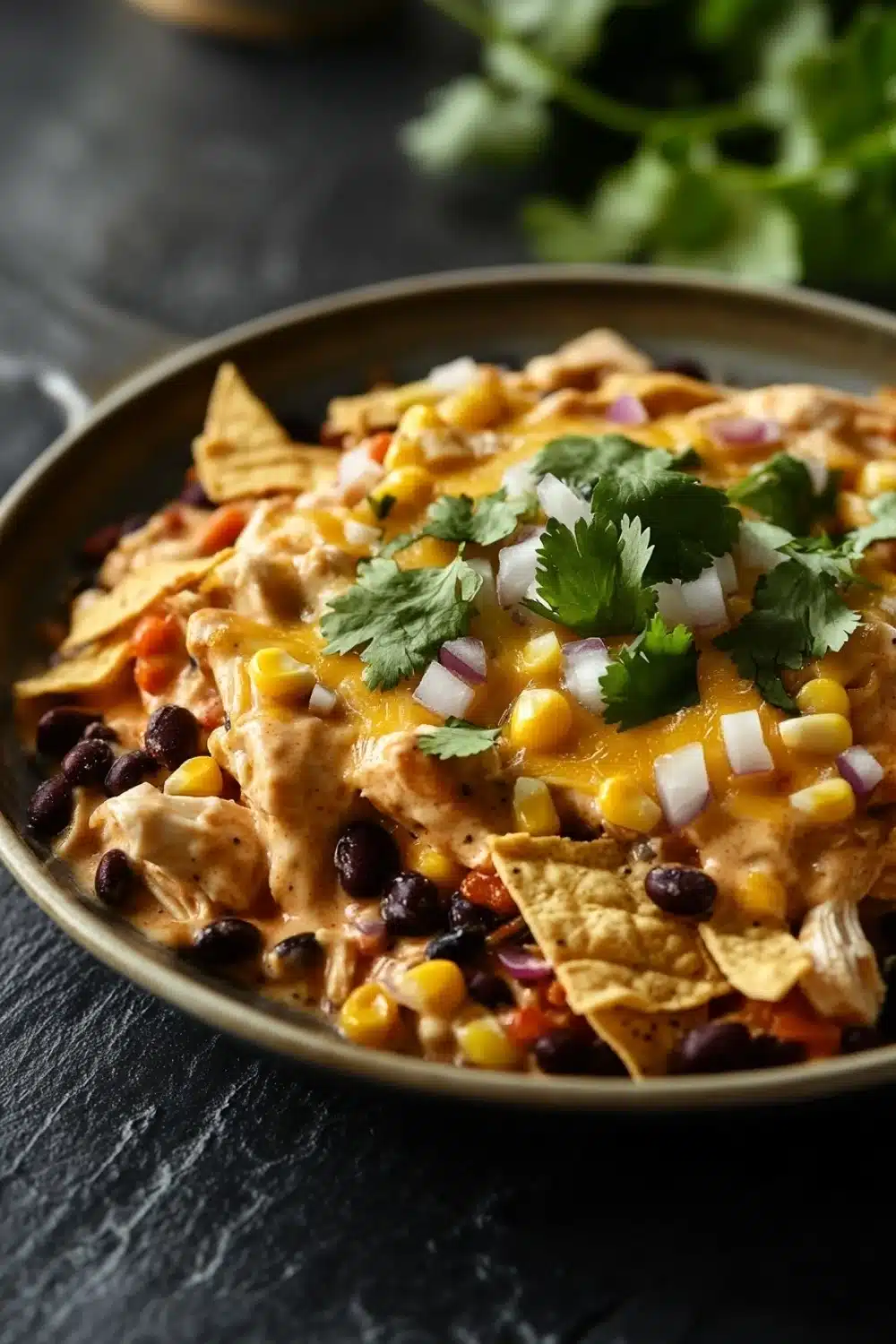 Maxis kitchen creamy chicken enchilada skillet - the image is a close-up of a bowl of nachos. the bowl is made of metal and is placed on a dark grey countertop. the nacho is made up of shredded chicken, corn kernels, black beans, red onions, and cilantro. the chicken is cooked to a golden brown color and appears to be seasoned with herbs and spices. there are also a few tortilla chips scattered around the bowl. in the background, there is a sprig of fresh cilantro and a small bowl of sauce. the overall color palette of the image is warm and inviting.