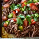 Mexican slow cooker beef roast - the image shows a rectangular baking tray filled with shredded beef and vegetables. the beef appears to be cooked medium-rare and is covered in a thick layer of melted cheese. the vegetables include diced tomatoes, red onions, and green jalapeno peppers. there are also some cilantro leaves scattered throughout the dish. the tray is sitting on a wooden table.