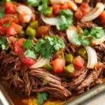 Mexican slow cooker beef roast - the image shows a close-up of a dish in a metal baking dish. the dish appears to be a mexican-style dish with shredded beef, diced tomatoes, onions, and green bell peppers. the beef is cooked medium-rare and is covered in a dark brown sauce. the tomatoes are bright red and appear to be seasoned with herbs and spices. the onions are sliced and are arranged in a pile on top of the beef. there are also a few sprigs of cilantro scattered throughout the dish, adding a pop of green color. the overall appearance is fresh and appetizing.