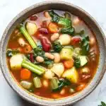 Minestrone soup italian - the image is a close-up of a bowl of soup on a white marble countertop. the soup appears to be a vegetable soup with chunks of white beans, carrots, celery, and other vegetables mixed in. the bowl is made of ceramic and has a brown rim. there are a few sprigs of parsley scattered around the bowl, adding a pop of green color to the dish. the background is blurred, making the bowl the focal point of the image.