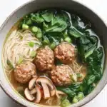 Molly yeh slow cooker asian wedding soup - the image is a close-up of a bowl of noodles and meatballs. the bowl is made of ceramic and has a light brown color. the noodles are white and appear to be thin and fluffy. the meatballs are brown and are arranged in a circular pattern on top of the noodles. there are also some sliced mushrooms and green onions scattered throughout the dish. the dish is garnished with fresh spinach leaves. the broth is a dark brown color and appears to be thick and creamy. the background is a white marble countertop.