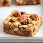 Monster oat cookie bar - the image shows a close-up of a square-shaped granola bar on a white baking tray. the bar is made with oats, nuts, and dried cranberries, and is topped with chocolate chips and pecans. the bars appear to be freshly baked and are arranged in a neat and orderly manner. the background is blurred, but it appears to be a kitchen countertop.