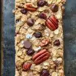 Monster oat cookie bar - the image shows a rectangular bar of granola on a baking tray. the granola appears to be made with oats, nuts, and dried cherries. there are also chunks of chocolate and pecans scattered throughout the granola. the bars are golden brown and look freshly baked. the tray is lined with parchment paper, and the background is dark.