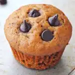 Muffin dolci allo yogurt e cioccolato - the image is of a freshly baked muffin on a white plate. the muffin is golden brown in color and has a crumbly texture. it is topped with chocolate chips, which are arranged in a circular pattern around the edges of the muffin. the plate is sitting on a wooden surface with a few scattered chocolate chips around it. the background is blurred, but it appears to be a kitchen countertop.