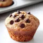 Muffin dolci allo yogurt e cioccolato - the image shows a freshly baked muffin in a muffin tin. the muffin is golden brown in color and has a crumbly texture. it is topped with chocolate chips, which are scattered across the top of the muffin. in the background, there are more muffins in the muffins, but they are slightly blurred. the tray is white and appears to be made of metal.