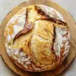 No knead bread dutch oven 2 hour - the image is a close-up of a freshly baked loaf of bread on a wooden cutting board. the bread appears to be freshly baked, with a golden-brown crust and a soft, fluffy interior. the edges of the bread are slightly frayed, indicating that it has been freshly baked. the cutting board is placed on a beige-colored surface, possibly a table or countertop. the background is blurred, making the bread the focal point of the image.