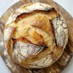 No knead bread dutch oven rise - the image shows a round loaf of bread on a wooden cutting board. the bread appears to be freshly baked and has a golden brown crust on top. the inside of the bread is visible, with a criss-cross pattern of brown and white swirls. the edges of the crust are slightly frayed, indicating that the bread has been freshly baked. the cutting board is placed on a white marble countertop.