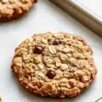 Oatmeal cookie lactation - the image shows two freshly baked oatmeal cookies on a baking tray. the cookies are golden brown and have a crumbly texture. they are topped with chopped nuts and chocolate chips. the tray is lined with parchment paper and there are two more cookies visible on the left side of the image. the background is blurred, but it appears to be a kitchen countertop.