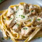 One pot chicken fettuccine - the image shows a plate of tagliatelle pasta with chicken and parmesan cheese. the pasta is cooked al dente and is topped with chunks of chicken and a creamy sauce. the dish is garnished with fresh parsley. the plate is sitting on a dark grey countertop with a few sprigs of parsley scattered around. the background is blurred, but it appears to be a kitchen countertop.