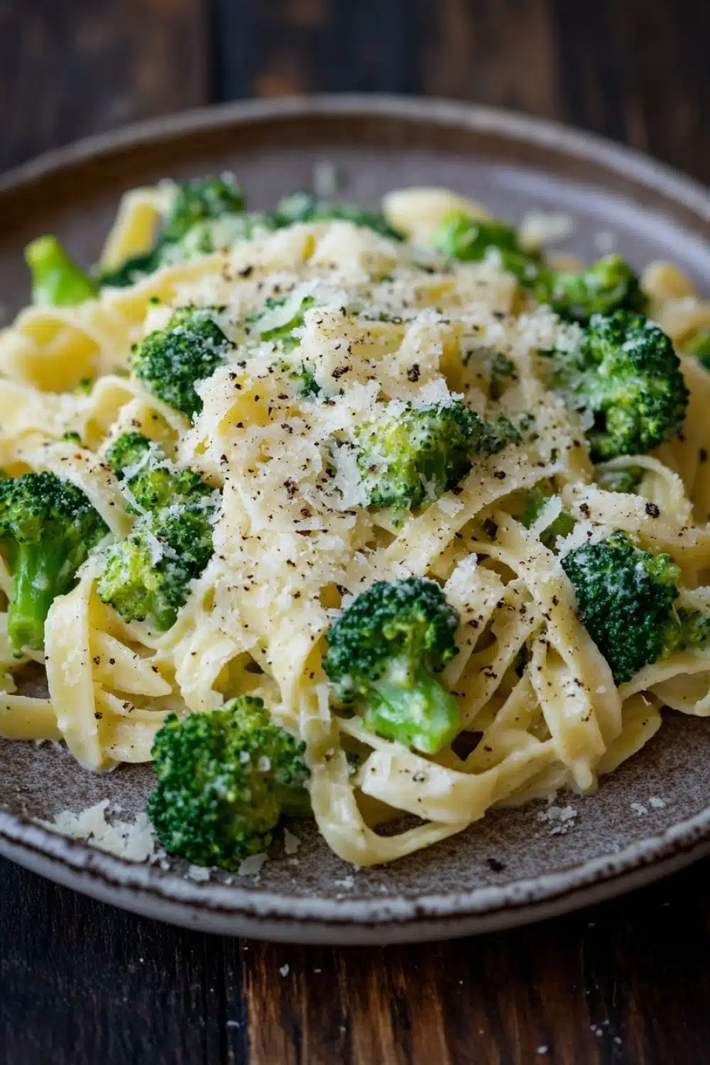One pot creamy broccoli and parmesan noodles - the image shows a plate of pasta dish with broccoli florets. the pasta is fettuccine alfredo and is topped with grated parmesan cheese. the dish is served on a brown plate with a wooden table in the background. the broccoli is bright green and appears to be cooked until it is soft and fluffy. the cheese is sprinkled on top, adding a creamy texture to the dish.