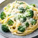 One pot creamy broccoli and parmesan noodles - the image shows a plate of fettuccine alfredo pasta with broccoli florets. the pasta is cooked al dente and is topped with a generous amount of parmesan cheese. the broccoli is bright green and appears to be fresh and healthy. the plate is gray with a speckled pattern and is sitting on a white marble countertop. the background is blurred, making the pasta the focal point of the image.