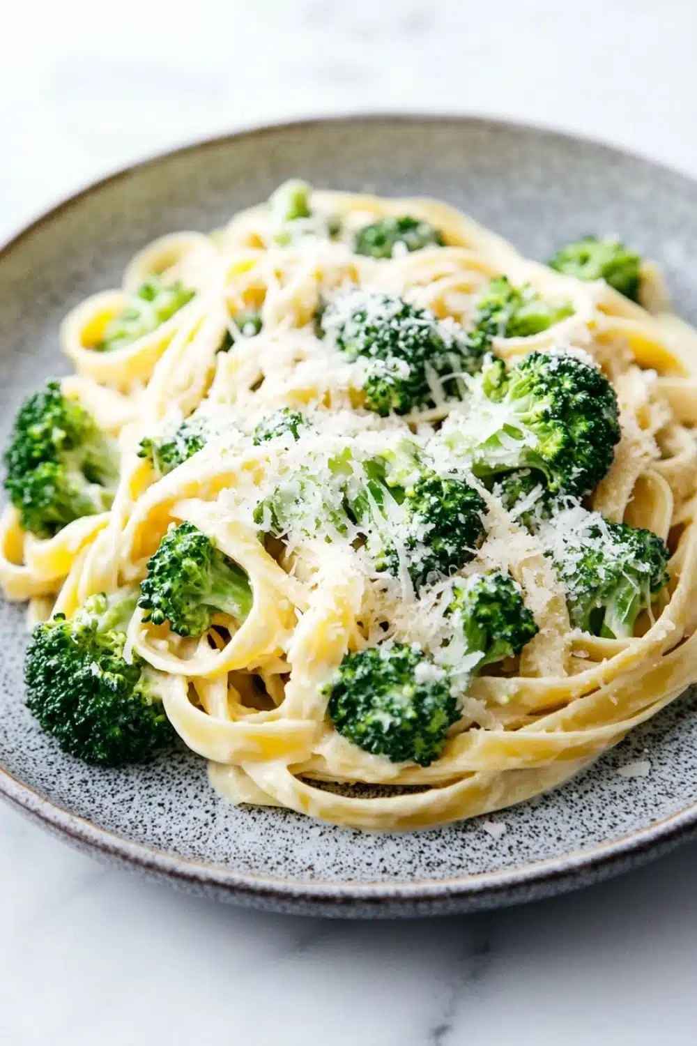 One pot creamy broccoli and parmesan noodles - the image shows a plate of fettuccine alfredo pasta with broccoli florets. the pasta is cooked al dente and is topped with a generous amount of parmesan cheese. the broccoli is bright green and appears to be fresh and healthy. the plate is gray with a speckled pattern and is sitting on a white marble countertop. the background is blurred, making the pasta the focal point of the image.