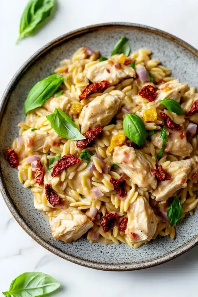 One pot creamy sun dried tomato chicken and orzo - the image is a close-up of a bowl of pasta dish. the pasta appears to be fusilli, which is a type of spiral-shaped pasta, and is topped with chunks of chicken, sun-dried tomatoes, red onions, and basil leaves. the dish is garnished with fresh basil leaves, giving it a fresh and vibrant look. the bowl is sitting on a white marble countertop, and there are a few basil leaves scattered around the bowl. the overall color palette of the dish is warm and inviting, with shades of yellow, orange, and red.