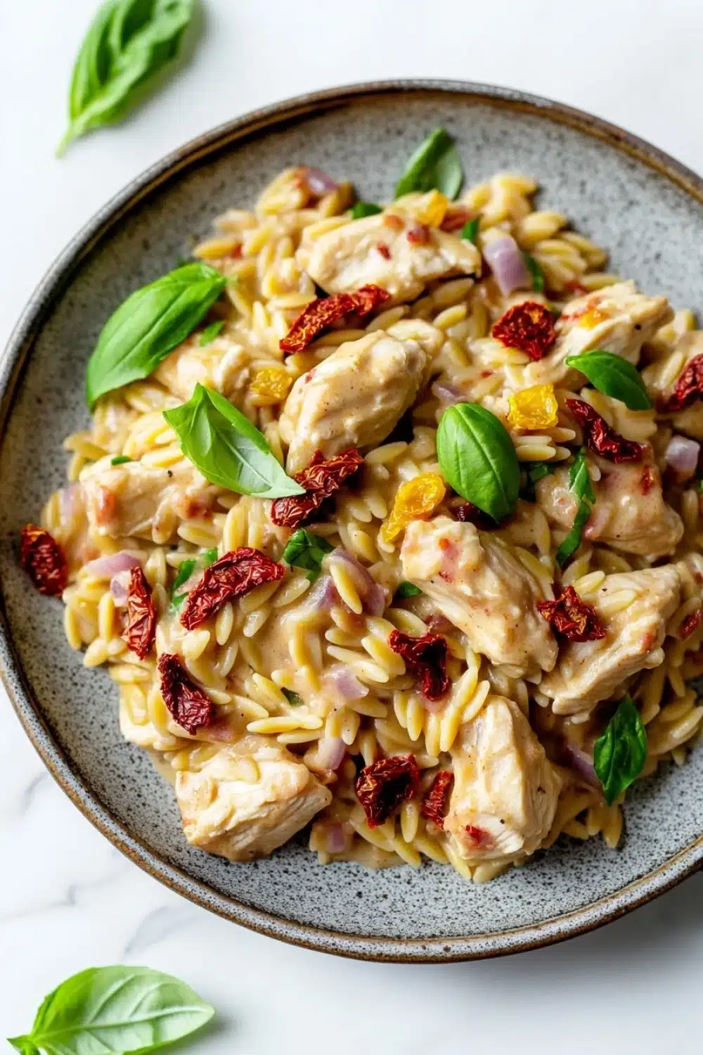 One pot creamy sun dried tomato chicken and orzo - the image is a close-up of a bowl of pasta dish. the pasta appears to be fusilli, which is a type of spiral-shaped pasta, and is topped with chunks of chicken, sun-dried tomatoes, red onions, and basil leaves. the dish is garnished with fresh basil leaves, giving it a fresh and vibrant look. the bowl is sitting on a white marble countertop, and there are a few basil leaves scattered around the bowl. the overall color palette of the dish is warm and inviting, with shades of yellow, orange, and red.