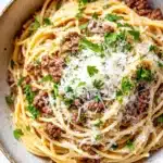 One pot garlic butter pasta with beef - the image is a close-up of a bowl of spaghetti bolognese. the spaghetti is cooked al dente and is topped with a generous amount of minced meat and grated parmesan cheese. the dish is garnished with fresh parsley. the bowl is made of ceramic and is sitting on a white marble countertop. the background is blurred, making the spaghetti the focal point of the image.