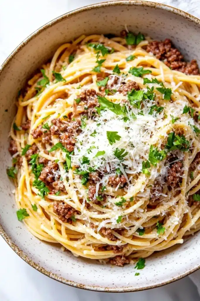 One pot garlic butter pasta with beef - the image is a close-up of a bowl of spaghetti bolognese. the spaghetti is cooked al dente and is topped with a generous amount of minced meat and grated parmesan cheese. the dish is garnished with fresh parsley. the bowl is made of ceramic and is sitting on a white marble countertop. the background is blurred, making the spaghetti the focal point of the image.