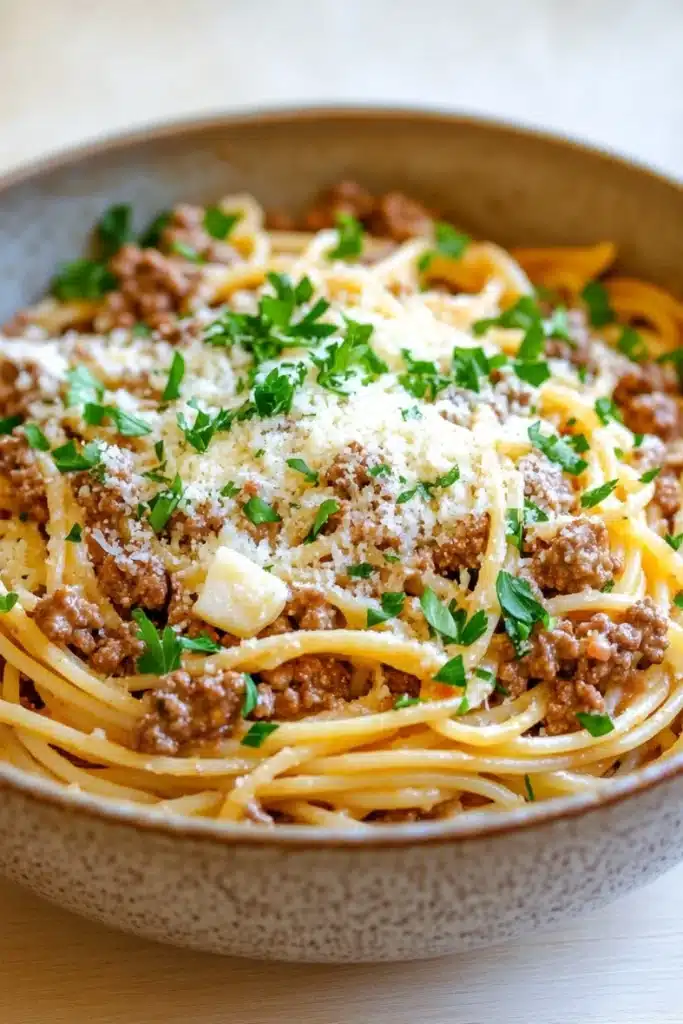One pot garlic butter pasta with beef - the image is a close-up of a bowl of spaghetti bolognese. the spaghetti is cooked al dente and is topped with a generous amount of minced meat and grated parmesan cheese. the dish is garnished with fresh parsley. the bowl is made of ceramic and is sitting on a wooden table. the background is blurred, but it appears to be a kitchen countertop.