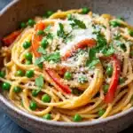 One pot pasta mit erbsen und paprika - the image is a close-up of a bowl of spaghetti dish. the bowl is made of ceramic and has a speckled pattern. the spaghetti is cooked al dente and is topped with sliced red bell peppers, green peas, and grated parmesan cheese. the dish is garnished with fresh parsley. the background is a dark grey countertop.