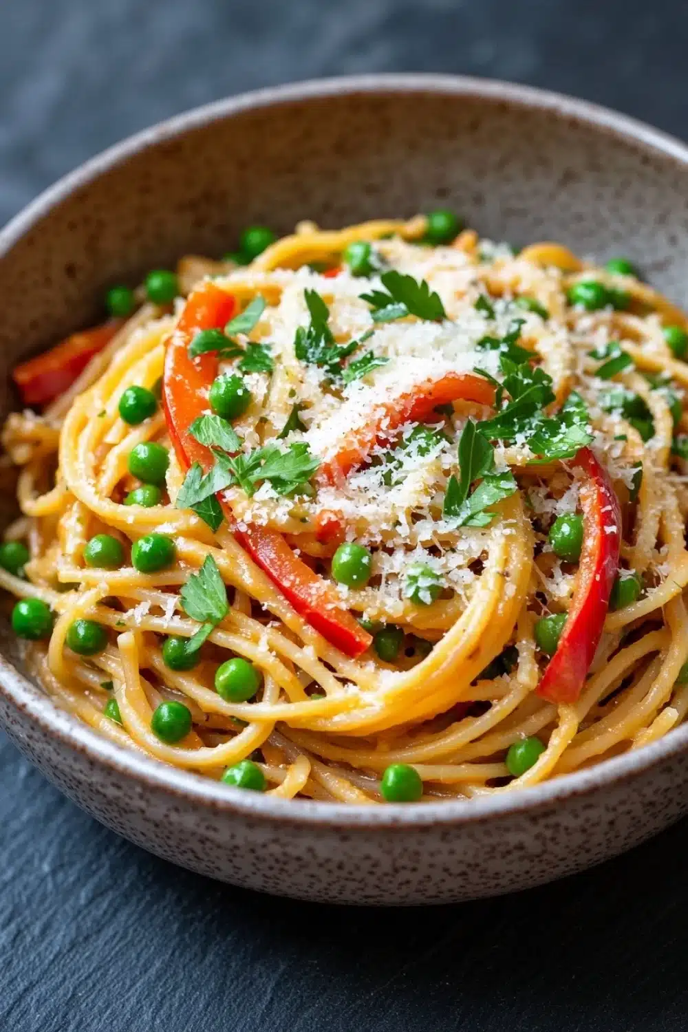One pot pasta mit erbsen und paprika - the image is a close-up of a bowl of spaghetti dish. the bowl is made of ceramic and has a speckled pattern. the spaghetti is cooked al dente and is topped with sliced red bell peppers, green peas, and grated parmesan cheese. the dish is garnished with fresh parsley. the background is a dark grey countertop.