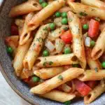 One pot pasta mit erbsen und paprika - the image is a close-up of a bowl of pasta dish. the pasta appears to be penne, which is a type of penne pasta, and it is cooked in a tomato-based sauce. the dish is garnished with chopped herbs, including parsley, and there are small pieces of red and green peas scattered throughout. the colors of the dish are vibrant and the texture looks fresh and appetizing. the bowl is made of ceramic and is sitting on a wooden table.