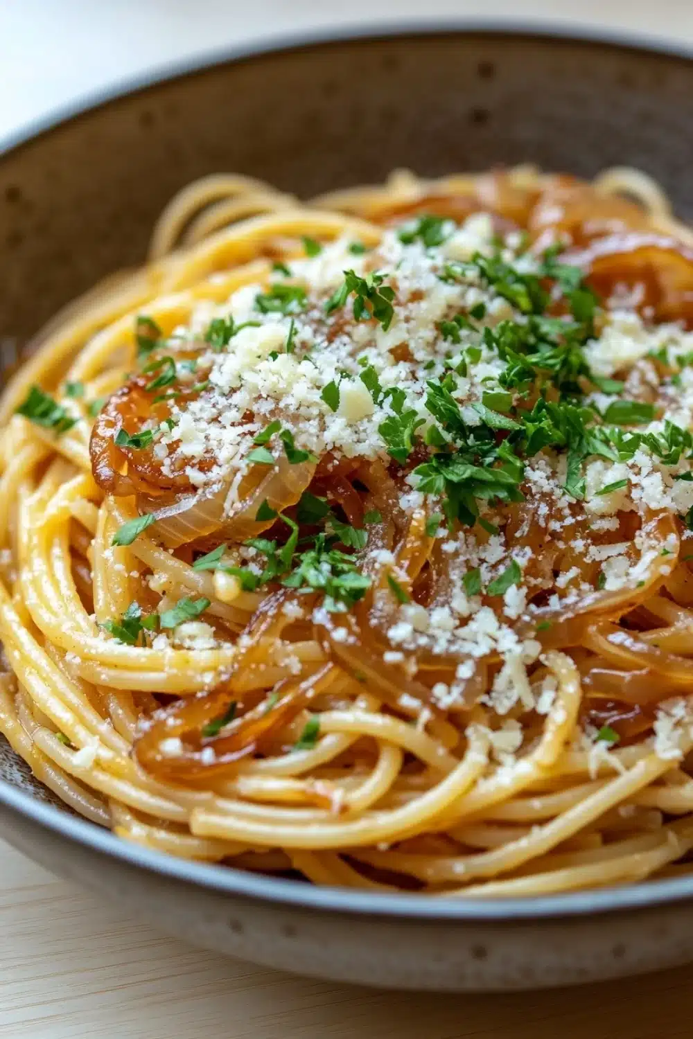One pot pasta zwiebel - the image is a close-up of a plate of spaghetti. the spaghetti is cooked al dente and is topped with a generous amount of grated parmesan cheese. the dish is garnished with fresh parsley and sliced onions. the plate is made of dark brown ceramic and is sitting on a wooden table. the background is blurred, but it appears to be a kitchen countertop.