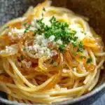 One pot pasta zwiebel - the image is a close-up of a bowl of spaghetti. the spaghetti is cooked al dente and is topped with sliced onions and grated parmesan cheese. the dish is garnished with a sprig of fresh parsley. the bowl is made of ceramic and has a dark brown color with a speckled pattern. the background is blurred, but it appears to be a kitchen countertop.