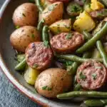 One pot sausage green beans potatoes - the image is a close-up of a plate of food. the plate is made of ceramic and is placed on a dark grey tablecloth. the food appears to be a salad with green beans, potatoes, and sausage. the potatoes are golden brown and appear to be seasoned with herbs and spices. the sausage is cut into small cubes and is arranged in a circular pattern on top of the green beans. the green beans are bright green and look fresh and vibrant. there are also small pieces of yellow bell peppers scattered throughout the salad. the overall appearance of the dish is colorful and appetizing.