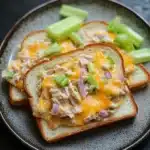 Openfaced tuna melt sandwich - the image shows a plate with three slices of bread on it. the bread is toasted and has a golden brown crust. on top of the bread, there is a mixture of diced chicken, diced onions, and green celery. the chicken appears to be cooked and is covered in a creamy yellow sauce. the celery is cut into small pieces and is scattered around the plate. the plate is black and the background is blurred, making the plate the focal point of the image.