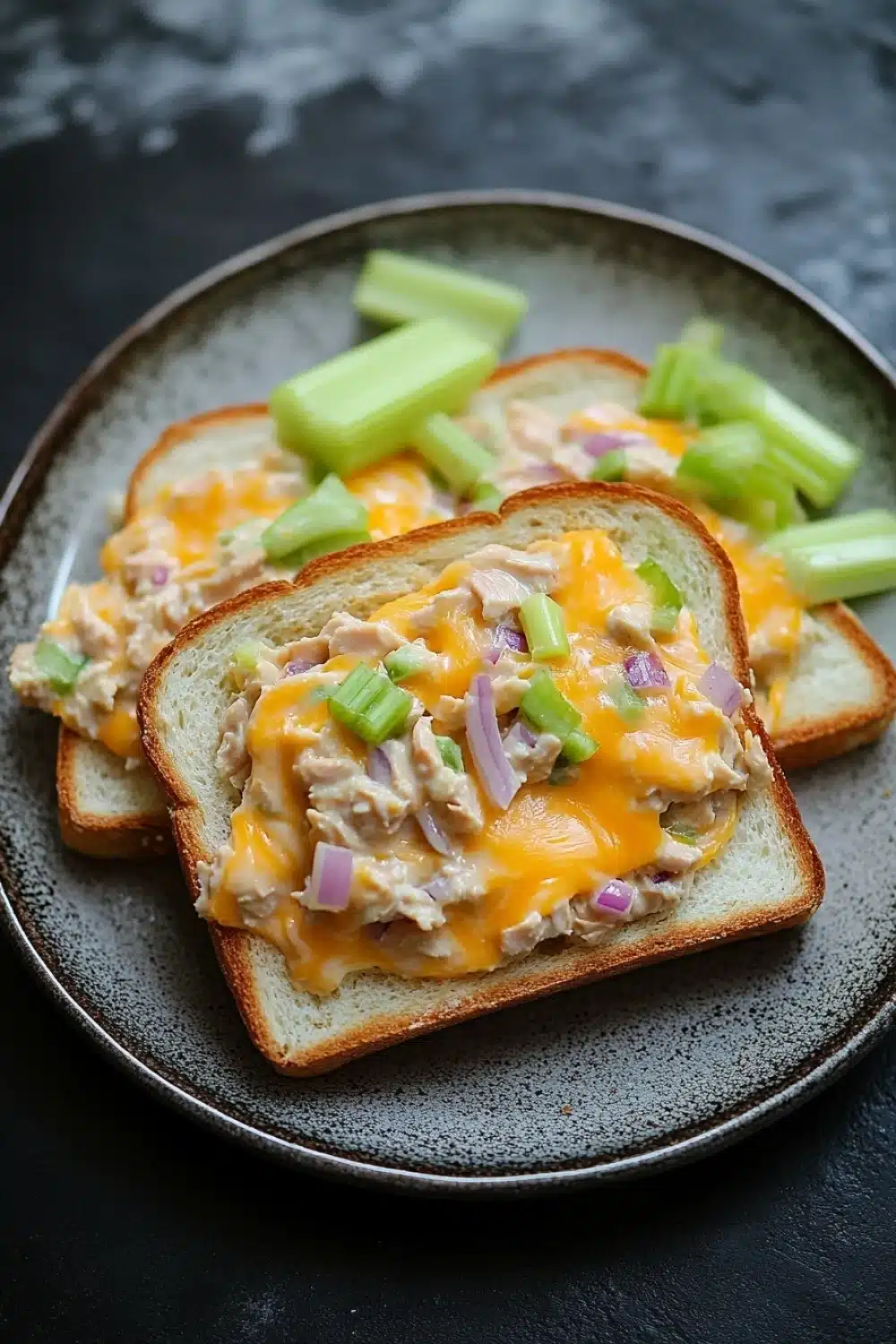 Openfaced tuna melt sandwich - the image shows a plate with three slices of bread on it. the bread is toasted and has a golden brown crust. on top of the bread, there is a mixture of diced chicken, diced onions, and green celery. the chicken appears to be cooked and is covered in a creamy yellow sauce. the celery is cut into small pieces and is scattered around the plate. the plate is black and the background is blurred, making the plate the focal point of the image.