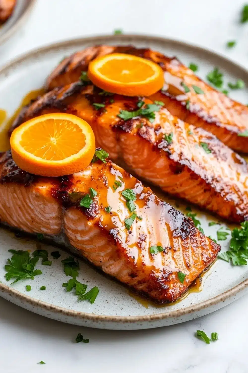 Orange glazed salmon air fryer - the image shows two pieces of salmon on a white plate. the salmon is cooked to a golden brown color and is garnished with fresh parsley. there are two slices of orange on top of the salmon, adding a pop of color to the dish. the plate is sitting on a marble countertop, and there is another plate visible in the background. the overall presentation of the dish is elegant and appetizing.