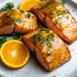 Orange glazed salmon air fryer - the image shows two pieces of salmon on a white plate with a blue and white speckled pattern. the salmon is cooked to a golden brown color and is garnished with fresh parsley. there are two slices of orange on the plate, one on each side of the salmon. the plate is sitting on a wooden table with a white background.
