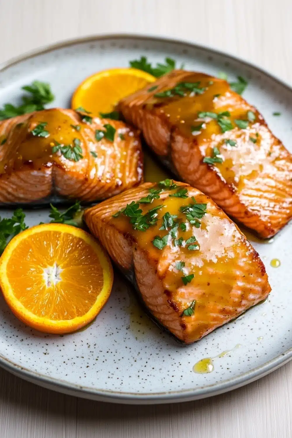 Orange glazed salmon air fryer - the image shows two pieces of salmon on a white plate with a blue and white speckled pattern. the salmon is cooked to a golden brown color and is garnished with fresh parsley. there are two slices of orange on the plate, one on each side of the salmon. the plate is sitting on a wooden table with a white background.