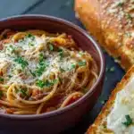 Pasta dinner garlic bread - the image shows a bowl of spaghetti with tomato sauce and parmesan cheese on top. the bowl is made of ceramic and is placed on a dark wooden table. next to the bowl, there is a slice of toasted bread with a golden brown crust. the bread appears to be freshly baked and has a crumbly texture. the dish is garnished with fresh herbs, adding a pop of green color to the dish. the background is blurred, but it seems to be a kitchen countertop.