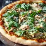 Pesto chicken arugula pizza - the image is of a freshly baked pizza on a wooden cutting board. the pizza has a golden brown crust and is topped with a generous amount of melted cheese and fresh green arugula leaves. the cheese appears to be melted and bubbly, and there are small pieces of chicken scattered throughout the pizza. the background is dark and out of focus, making the pizza the focal point of the image. there are a few sprigs of fresh herbs scattered around the cutting board, adding a pop of color to the scene.